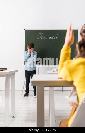 fuoco selettivo di scolari rovesciati che coprono il viso mentre si sta in piedi vicino a lavagna e compagno di classe con mano sollevata Foto Stock