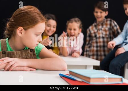 fuoco selettivo di scolari rovesciati seduti vicino ai compagni di classe che ride e che punta con le dita isolate su nero, concetto di bullismo Foto Stock