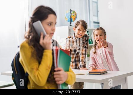 fuoco selettivo del compagno di classe crudele che punta con il dito contro la scolari turbato che parla sullo smartphone, concetto di bullismo Foto Stock