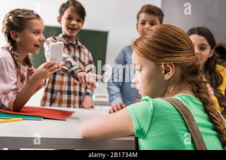 fuoco selettivo di scolaretta depressa seduta vicino a scholkid con lo smartphone e i classmates crudeli, concetto di cyberbullismo Foto Stock