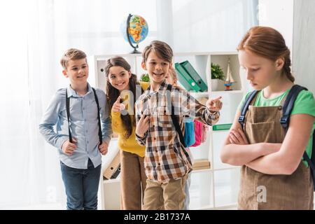 fuoco selettivo di scolari crudeli che puntano con le dita e ridendo vicino scolari bullied che si levano in piedi con le armi incrociate in classe Foto Stock