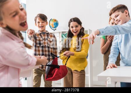 fuoco selettivo di allievi crudeli che tengono lo zaino e che punta con le dita al compagno di classe bullied urlando in classe Foto Stock