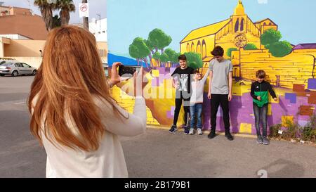 Madre che prende la foto dei suoi figli di fronte ad un muro con una penetura della cattedrale di Alcúdia, Spagna, Isole Baleari, Maiorca, Alcudia Foto Stock
