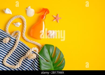 Lozione spray abbronzante in bottiglia d'arancia, borsa da spiaggia a righe, foglia tropicale e conchiglie su sfondo giallo. Vista dall'alto, piatto. Cosmesi estivi per la cura della pelle Foto Stock