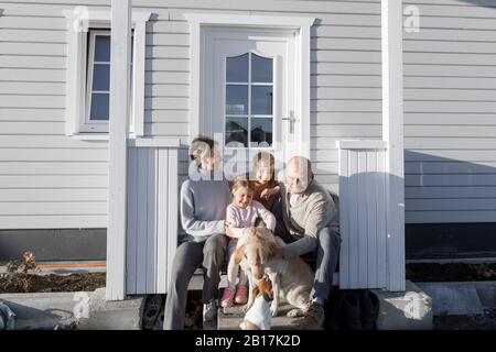 Felice famiglia con due bambini e due cani seduti di fronte alla loro casa Foto Stock