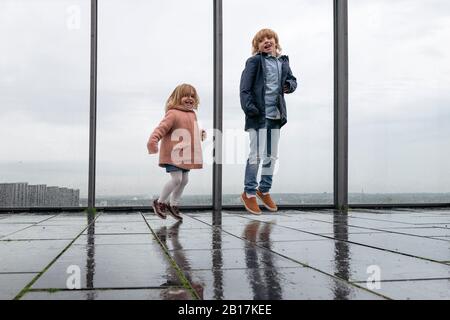 Fratello e sorella piccola che saltano sulla terrazza con vista il giorno delle piogge Foto Stock