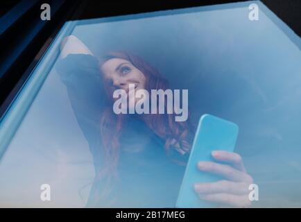 Ritratto di donna sorridente redheaded che tiene il telefono cellulare dietro il windowpane Foto Stock