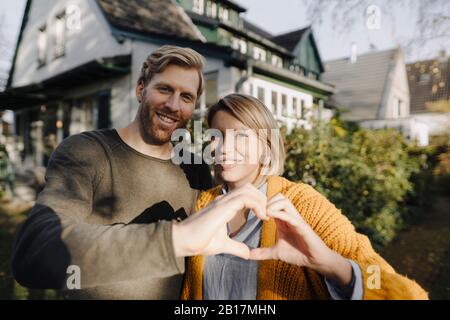 Ritratto di coppia sorridente in piedi davanti alla loro casa che forma un cuore con le loro mani Foto Stock