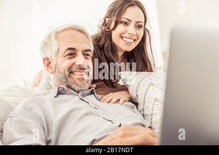 Uomo maturo sorridente che usa il laptop sul divano a casa con la donna che guarda Foto Stock