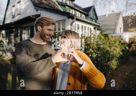 Coppia sorridente in piedi davanti alla loro casa che forma un cuore con le loro mani Foto Stock