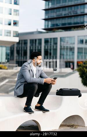 Giovane uomo d'affari informale seduto su una panchina della città utilizzando il telefono cellulare Foto Stock