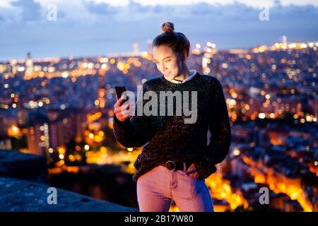Giovane donna seduta sulla ringhiera di protezione al di sopra della città utilizzando il telefono cellulare, Barcellona, Spagna Foto Stock