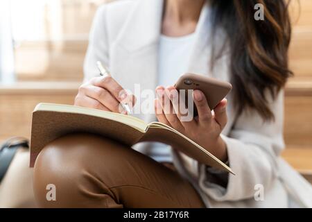 Primo piano di una donna seduta sulle scale utilizzando lo smartphone e prendendo appunti Foto Stock