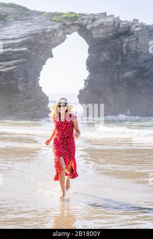 Donna bionda con abito rosso e cappello e corsa in spiaggia, Arco Naturale a Playa de Las Catedrales, Spagna Foto Stock