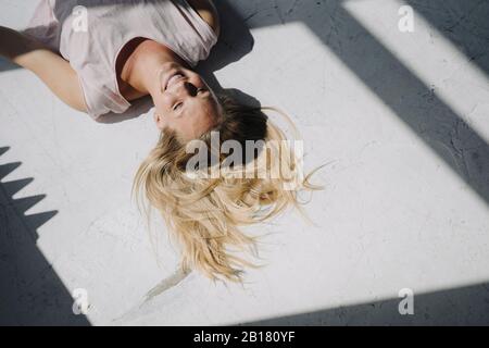 Giovane donna bionda sorridente sdraiata sul pavimento alla luce del sole Foto Stock