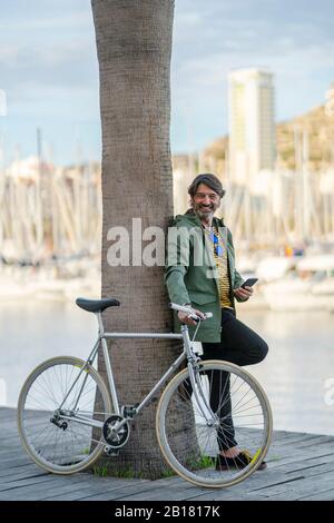 Ritratto di uomo maturo rilassato con bici fixie appoggiata contro il tronco di palma, Alicante, Spagna Foto Stock