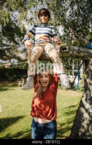 Ragazzo che arrampica l'albero, seduto sul ramo, madre che tiene le mani Foto Stock
