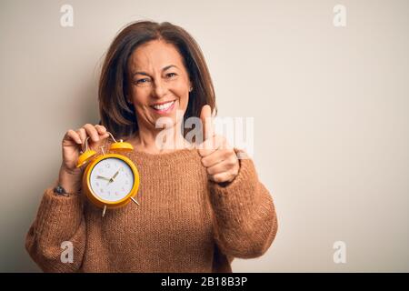 Donna di media età bruna che tiene l'orologio clasico di allarme sopra lo sfondo isolato che approva facendo il gesto positivo con la mano, pollici in su sorridente e felice Foto Stock