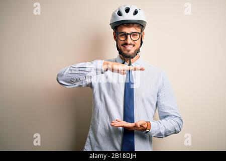 Giovane uomo d'affari che indossa occhiali e casco da bici in piedi su isolato bianco panbakground gesturing con le mani mostrando grande e grande segno, misura Foto Stock