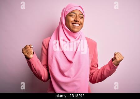 Giovane afro afroamericana indossando hijab musulmano su sfondo rosa isolato molto felice ed eccitato facendo il gesto del vincitore con le braccia sollevate, smi Foto Stock