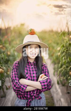 Ragazza asiatica carina con pomodoro rosso su cappello , raccogliendo verdure fresche in giardino Foto Stock