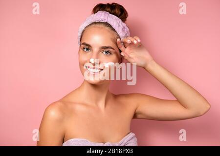 Una ragazza con la pelle pulita si alza su uno sfondo rosa, sorride alla fotocamera, regolando il bendaggio sulla testa, con la crema sul viso Foto Stock