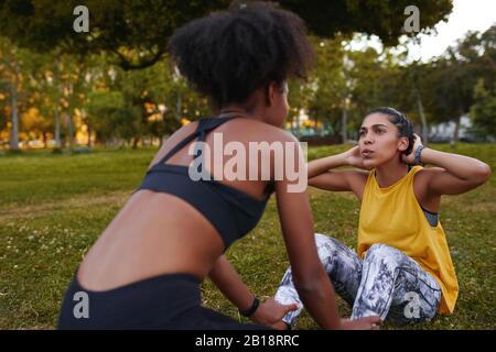 Adatta giovane donna che fa crunches con il suo compagno di esercitazione femmina sull'erba verde durante una sessione di workout - giovane donna determinata e motivata che fa Foto Stock