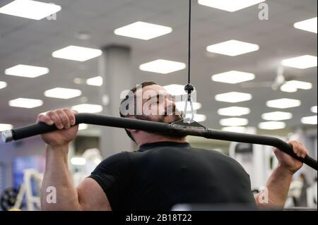 giovane uomo con bearded in maglia nera che fa l'esercitazione nella palestra interna di sport con l'apparecchiatura di idoneità Foto Stock