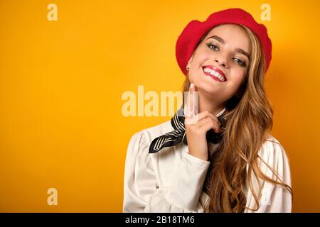 Una ragazza in un beretto rosso è in piedi su uno sfondo giallo, la testa è buttata indietro sorridendo, con un dito al mento Foto Stock