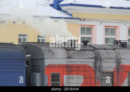 Fumo o vapore proviene da un camino sul tetto di una carrozza ferroviaria in inverno presso una stazione ferroviaria in Russia contro un edificio giallo. Foto Stock