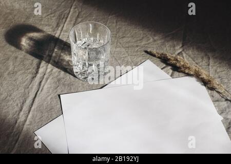 Cancelleria ancora vita scena. Vetro di acqua, erba asciutta su fondo di tela di lino beige del tavolo in luce del sole. Fogli di carta vuoti, carte mockup scena con Foto Stock