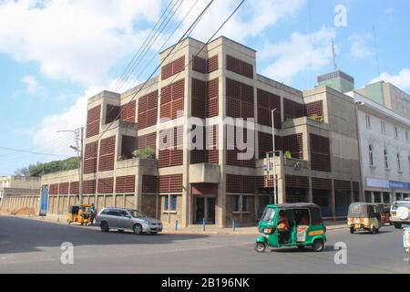 Mombasa, Kenya - 20 febbraio 2015: Strada ordinaria della città con un grande edificio nel bel mezzo della giornata. TukTuk si trova su una strada in Africa. Foto Stock