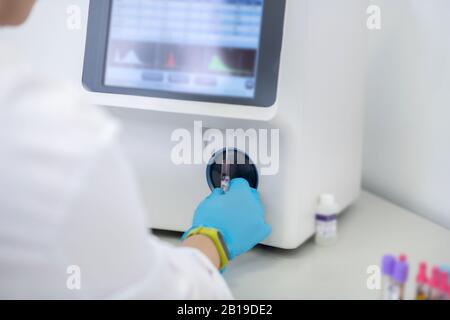 Tecnico in guanti blu premendo il pulsante di avvio dell'analizzatore ematologico in laboratorio Foto Stock