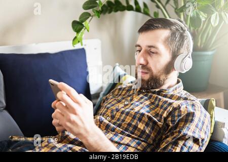Uomo che ascolta la musica con le cuffie sul divano di casa, guardando lo schermo del telefono cellulare Foto Stock