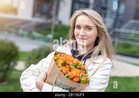 Ritratto giovane adulto sorridente elegante donna modello mano che tiene autentico fresco arancio spray rose fiore bouquet avvolto in carta artigianale sacchetto sulla città Foto Stock