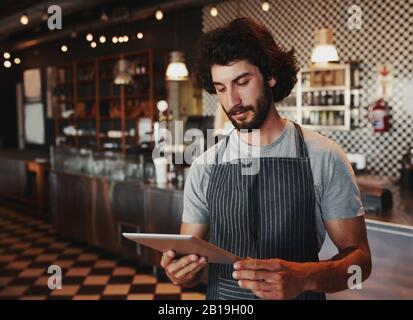 Giovane proprietario di caffè che controlla l'ordine in linea sul tablet digitale mentre in piedi nella casa del caffè che indossa grembiule nero Foto Stock