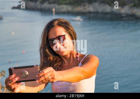 Donna che si rilassa al mare facendo selfie al tramonto, oceano sullo sfondo, vista frontale. Foto Stock
