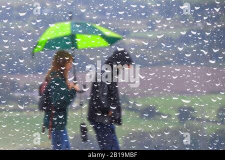 Riassunto di una coppia che cammina sotto la pioggia con un ombrello, visto attraverso una finestra coperta di goccioline di pioggia. Wet tempo UK. Foto Stock
