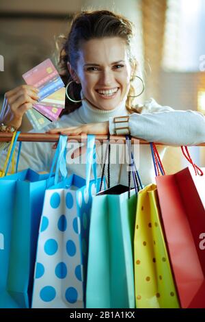 Ritratto di donna sorridente elegante di 40 anni in maglione bianco e gonna vicino a borse colorate appese su barra di rame vestiti con un ventilatore di cre Foto Stock