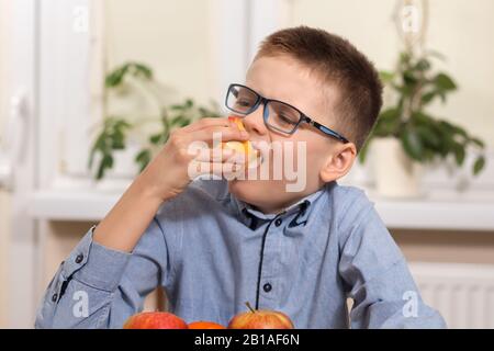 Il ragazzo in una camicia blu e bicchieri si siede al tavolo e tiene in mano una mela che mangia con gusto. Foto Stock