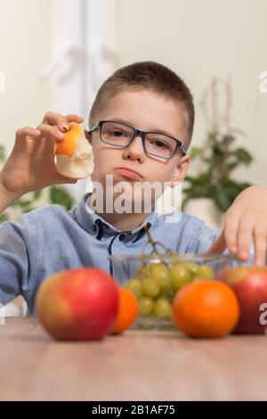 Il ragazzo in una camicia blu e bicchieri si siede al tavolo e tiene in mano una mela che mangia con gusto. Foto Stock