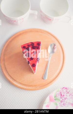 Fetta di cheesecake di ribes e cucchiaio su tavola rotonda di legno. Due tazze per tè, piattino su tavola bianca. Torta al formaggio con frutti di bosco. Vista dall'alto Foto Stock