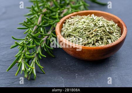 Rametti di rosmarino freschi e una ciotola di legno con rosmarino essiccato su uno sfondo di ardesia nera Foto Stock