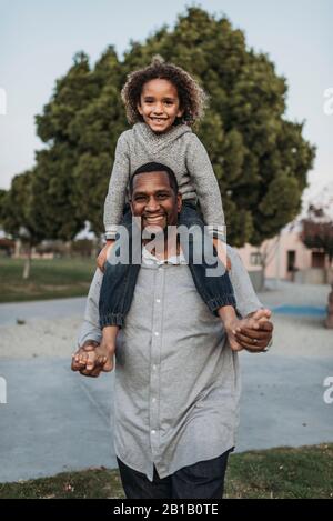 Un figlio gioioso seduto sulle spalle di un padre felice al parco giochi Foto Stock