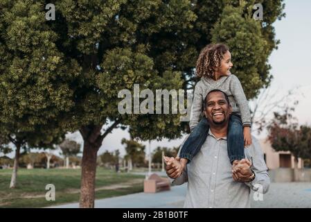 Un figlio gioioso seduto sulle spalle di un padre felice al parco giochi Foto Stock
