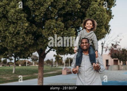 Un figlio gioioso seduto sulle spalle di un padre felice al parco giochi Foto Stock