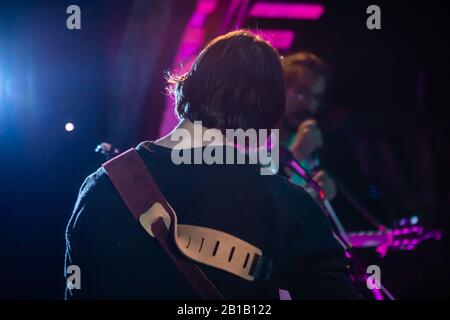 Un closeup soft focus vista da dietro un musicista che si esibisce sul palco con i membri della band blurry, in background e viola scena luce di notte al festival Foto Stock