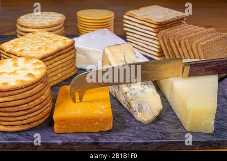 Mucchi di cracker cotti a secco e diversi tipi di formaggio e un coltello su un tagliere di granito. Foto Stock