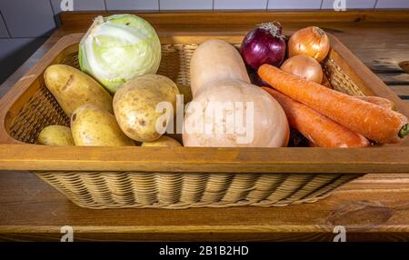 Un cestino di vimini su un piano di lavoro della cucina / contatore che conteggia le verdure di radice crude - cavolo, patate, carote, zucca del butternut e cipolle. Foto Stock