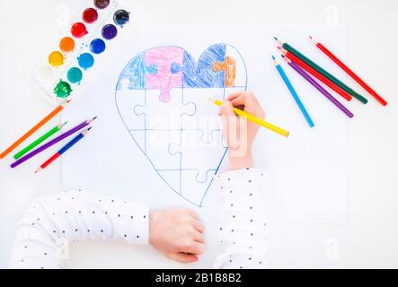 Un bambino sta disegnando cuore colorato con puzzle jigsaw su sfondo bianco come un simbolo di autismo, con matite colorate e acquerelli. Foto Stock